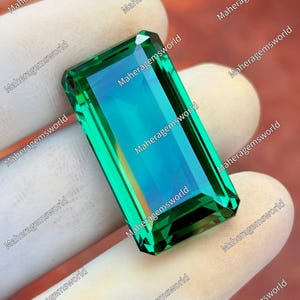 May include: A rectangular, emerald-cut gemstone with a vibrant green hue and a hint of blue. The gem is held by a gloved hand, showcasing its facets and clarity. The text "Maheragemsworld" is repeated around the edges of the image.