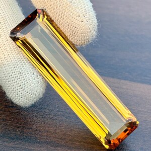 Könnte beinhalten: Ein rechteckiger, facettierter Edelstein in goldgelber Farbe mit bernsteinfarbenen Kanten. Der Stein wird von einem weißen Handschuh gehalten, der seine Klarheit und seinen Schliff zeigt. Die Form des Edelsteins ist länglich.