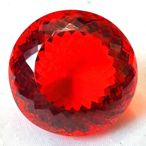 Può includere: Una gemma rossa, rotonda e sfaccettata, con taglio brillante. La gemma presenta un motivo centrale a forma di stella e riflette la luce, mettendo in risalto il suo design complesso. La gemma è di un rosso vibrante.