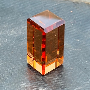 May include: A rectangular, amber-colored crystal sculpture with a faceted design. The transparent material allows light to pass through, highlighting the internal structure and creating a play of light and shadow. The sculpture has a geometric shape and a polished finish.