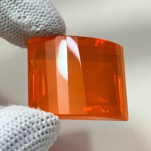 May include: A faceted, square-cut, orange gemstone held by a gloved hand. The gemstone has a rectangular shape with a curved top edge and multiple facets that reflect light. The color is a vibrant, translucent orange.