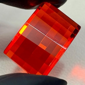 May include: A rectangular, faceted gemstone in a vivid red colour. The surface is divided into many small, square facets, giving a mosaic effect. Light refracts through the gem, highlighting its internal structure and colour variations. A striking piece.