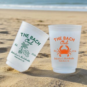 May include: Two white plastic cups on a sandy beach. One cup has green text that reads "THE BACH Club" with a palm tree graphic and "Sebastian's Bachelorette Malibu Beach, CA 2026." The other cup has orange text and a crab graphic.
