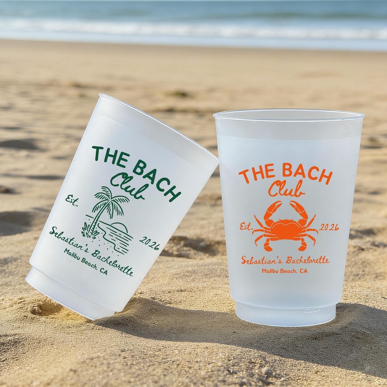 May include: Two white plastic cups on a sandy beach. One cup has green text that reads "THE BACH Club" with a palm tree graphic and "Sebastian's Bachelorette Malibu Beach, CA 2026." The other cup has orange text and a crab graphic.