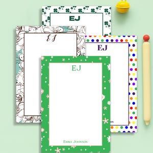 May include: A collection of personalized stationery pads with various border designs, including floral, polka dot, and shamrock patterns. Each pad features the initials "EJ" and some include a name. A yellow bell and a pen are also in the image.