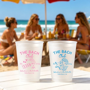 May include: Two white plastic cups with the text "THE BACH Club" in pink and blue, with beach-themed graphics. The cups are for Sebastian's Bachelorette in Malibu Beach, CA, with the year 2026. The background shows a beach scene.