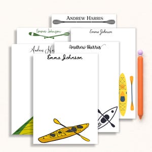 May include: A collection of white notepads with kayak and paddle illustrations in yellow, green, and black. The notepads feature the names "Andrew Harris" and "Emma Johnson" in various fonts. An orange pen with a purple tip is also present.