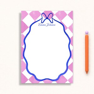 May include: A personalized notepad with a pink and white diamond pattern background. The notepad has a blue scalloped border and a blue bow at the top with the name "Emma Johnson" in script. An orange pen with a purple tip is next to the notepad.