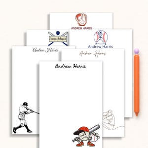 May include: A collection of white notepads with baseball-themed designs and the name "Andrew Harris" printed on them. The designs include a baseball glove, a baseball player, and a baseball with a bat. An orange pen is on the right.