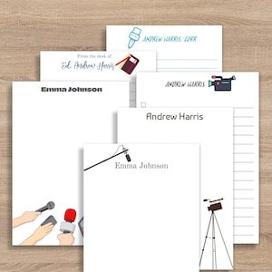 May include: A collection of white notepads with various designs and text. Some pads feature the names "Emma Johnson" and "Andrew Harris." Illustrations of a microphone, camera, and news-related icons are also present. The pads are arranged on a light wood surface.
