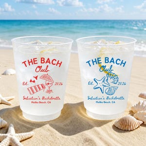 May include: Two clear plastic cups filled with a beverage and ice, with the text "THE BACH Club" in red and blue. The cups are decorated with beach-themed graphics and the text "Sebastian's Bachelorette Malibu Beach, CA". The cups are on a sandy beach.