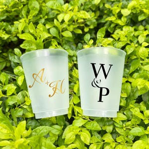 May include: Two frosted plastic cups with custom initials. One cup has gold initials "A + H", the other has black initials "W & P". The cups are set against a backdrop of green foliage.