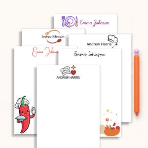May include: A collection of white notepads with various culinary-themed designs and text. Designs include a chef's hat, utensils, and a cartoon chili pepper. The names "Emma Johnson" and "Andrew Harris" are printed on some pads. An orange pen is on the right.