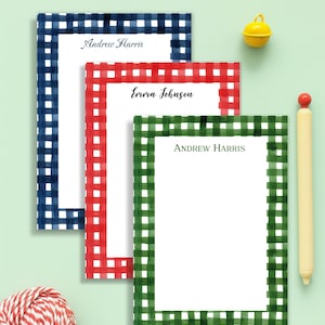 May include: Three personalized notepads with gingham borders in navy blue, red, and green. Each notepad has a name printed at the top: "Andrew Harris" and "Emma Johnson". A yellow bell and a pen are also in the image.