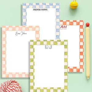 May include: Four personalized notepads with checkered borders in pink, blue, orange, and green. Each notepad has a name or initials printed at the top. A yellow bell and a pen are also visible.