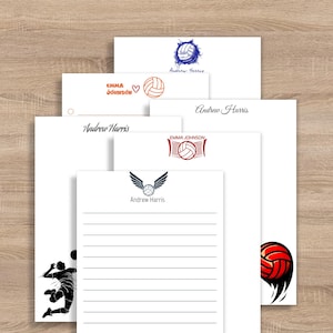 May include: A collection of white stationery featuring volleyball-themed designs. The designs include volleyballs, nets, and wings, with names like "Andrew Harris" and "Emma Johnson" printed on the stationery. The stationery is arranged on a wooden surface.