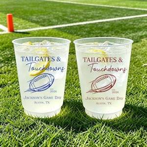 May include: Two clear plastic cups filled with ice and lemon slices. Each cup features the text TAILGATES & TOUCHDOWNS, a football graphic, EST. 2026, Jackson's Game Day, and Austin, TX. One cup has blue text and graphics, the other has maroon.