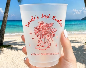 May include: A frosted plastic cup with red text and graphics. The text reads "Bride's Last Rodeo" with a floral and cowboy boot design. Below the design is the text "Olivia | Nashville 2026". The cup is held in front of a beach scene.