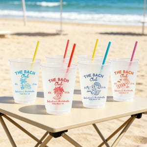 May include: Clear plastic cups with colorful straws are arranged on a table at the beach. Each cup is printed with "THE BACH Club" and a unique graphic, along with "Sebastian's Bachelorette" and the year 2026.