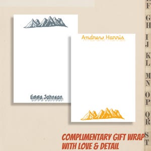 May include: Two white notepads with mountain illustrations. One notepad has a gray mountain design at the top and the name "Emma Johnson" at the bottom. The other notepad has an orange mountain design and the name "Andrew Harris".