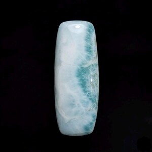 May include: A polished, elongated gemstone with a light blue and white color pattern, featuring areas of turquoise blue. The stone has a smooth, glossy surface and is set against a black background. The gemstone's shape is rectangular with rounded edges.