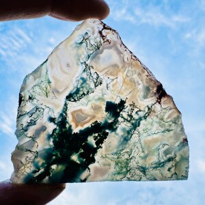 Può includere: Una pietra traslucida di forma irregolare, con un motivo naturale di vortici e venature verdi, marroni e bianche. La pietra è tenuta contro un cielo blu, mostrando i suoi intricati dettagli e la sua bellezza naturale. I bordi della pietra sono ruvidi e irregolari.