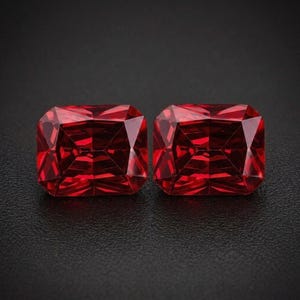 May include: Two radiant-cut, deep red gemstones. Each gem has a rectangular shape with faceted surfaces that catch the light, creating a brilliant display. The gems are set against a dark background, highlighting their rich color and clarity.