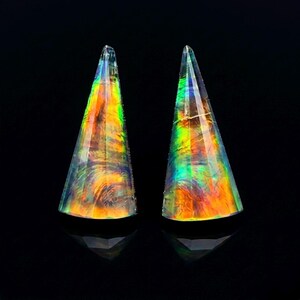 May include: Two faceted, cone-shaped objects with a translucent, iridescent finish. The surfaces display a vibrant spectrum of colors, including orange, yellow, green, and blue, which shift in intensity across the geometric, multi-faceted structure.