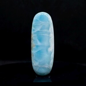 Puede incluir: Una gema pulida y alargada con una parte superior redondeada. La piedra presenta un color azul claro con vetas blancas y grises. La gema se muestra sobre un fondo negro, resaltando su superficie lisa y reflectante.