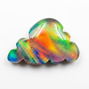 Pode incluir: Uma pedra preciosa polida em forma de nuvem que exibe um jogo de cores vibrante com flashes iridescentes de laranja, amarelo, verde e azul. A superfície é lisa e reflexiva, mostrando um espectro de tons variáveis em sua forma orgânica.