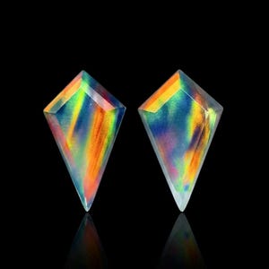 May include: Two kite-shaped gemstones featuring a vibrant iridescent play of color with streaks of orange, yellow, blue, and green. The stones have a smooth, polished surface and a pointed bottom edge, displaying a reflective, multi-colored sheen.