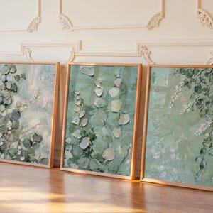 May include: Three framed abstract paintings with botanical themes. Each artwork features a textured, impasto style with shades of green and white, depicting leaves and floral elements. The frames are made of light-coloured wood, and the paintings are displayed on a wooden floor.