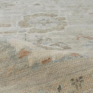 May include: Close-up of a light-colored rug with a faded floral pattern. The rug has a textured surface with a mix of beige, light blue, and rust-colored accents. The edges of the rug are slightly curled, revealing the woven texture.