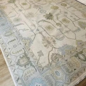 May include: A large, rectangular area rug with a floral pattern in shades of cream, gray, and sage green. The rug features a detailed border and a central design of stylized flowers and vines. The rug is placed on a wooden floor.