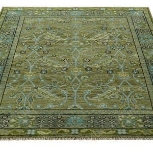 May include: A square, olive green rug with a detailed floral pattern and a fringe border. The rug features a central design with light blue accents and a darker green border, creating a visually appealing contrast. The rug's dimensions are not visible.