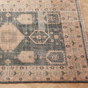 Peut inclure: Un tapis de sol d'aspect vieilli avec un centre gris foncé et une bordure couleur pêche. Le tapis présente des motifs géométriques et floraux en pêche et gris, avec une bordure à franges. Le tapis est sur un plancher en bois.