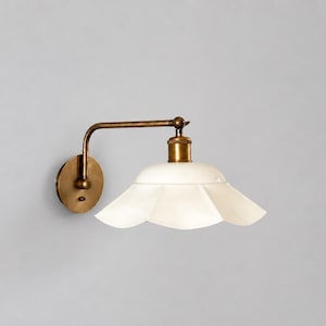 Puede incluir: Una lámpara de pared con acabado de latón y una pantalla blanca en forma de pétalo. La lámpara tiene un brazo curvo y una base redonda, creando una estética vintage. Diseñada para montaje en pared.
