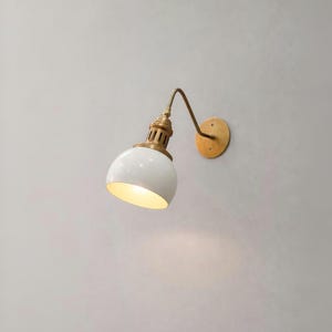 May include: A white and brass wall-mounted lamp. The lamp features a curved brass arm, a white, rounded shade, and a brass base. The lamp is mounted on a circular brass plate. The shade is approximately 15 cm in diameter.