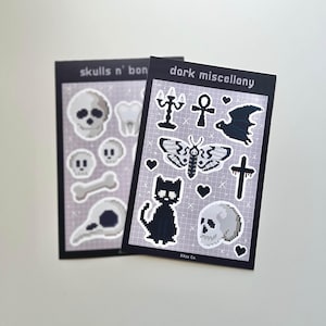 May include: Two sticker sheets with a dark theme. One sheet says "skulls n' bon" and features pixelated skulls, bones, and teeth. The other says "dark miscellany" and has a bat, moth, cat, cross, and other gothic-inspired designs.