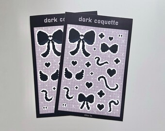 May include: Two sticker sheets with a dark coquette theme. Each sheet features pixelated black bows, hearts, and other shapes on a light purple background with a grid pattern. The text "dark coquette" is at the top of each sheet.