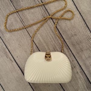 Puede incluir: Un bolso blanco estructurado con una correa de cadena dorada. El bolso tiene un diseño acanalado en forma de abanico en la parte delantera y un cierre dorado. La cadena es de estilo cuerda trenzada. El bolso está sobre una superficie de madera.