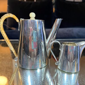May include: A silver tea set with a teapot and creamer. The teapot has a woven handle and a small, round knob on the lid. The creamer has a curved handle and spout. The set is displayed on a reflective surface.