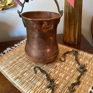 May include: A copper bucket with a brass handle and chain. The bucket has a worn, aged look and is sitting on a woven mat.