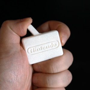 May include: A white, textured Nintendo-branded accessory. The rectangular piece has a small handle and the Nintendo logo is embossed on the front. The item is held in a hand against a black background.