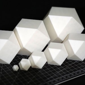 Puede incluir: Un conjunto de formas geométricas blancas de varios tamaños, dispuestas sobre una esterilla de corte negra. Las formas tienen múltiples facetas, creando una apariencia poligonal. La forma más grande mide unos 15 cm de ancho.