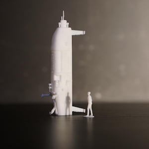 Peut inclure: Modèle blanc imprimé en 3D d'un sous-marin, avec des structures détaillées et des éléments en saillie. Une petite figurine se tient à côté du modèle, pour l'échelle. Le modèle de sous-marin est placé sur un fond sombre.