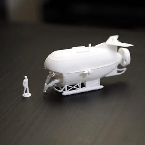 Peut inclure: Modèle blanc imprimé en 3D d'un zeppelin avec des détails complexes, dont une hélice et une structure de support. Une petite figurine se tient à côté du zeppelin, pour l'échelle. Le modèle est posé sur un fond sombre.