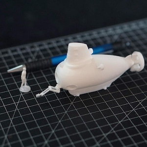 Peut inclure: Maquette de sous-marin blanc imprimée en 3D, avec une petite figurine et un stylo sur une surface quadrillée noire. Le sous-marin a une tour cylindrique et un corps arrondi avec de petits détails. La figurine est sur une base circulaire.