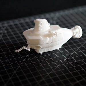 Peut inclure: Un modèle blanc de sous-marin avec un design détaillé, comprenant une tour de contrôle, des ailerons latéraux et une hélice arrière. Le sous-marin miniature repose sur une surface de grille noire.