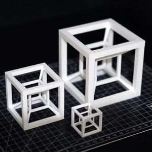 May include: Three white geometric cube sculptures of varying sizes are arranged on a black cutting mat. The cubes have an open, nested design, creating an optical illusion. The cutting mat has a grid pattern and measurement markings.